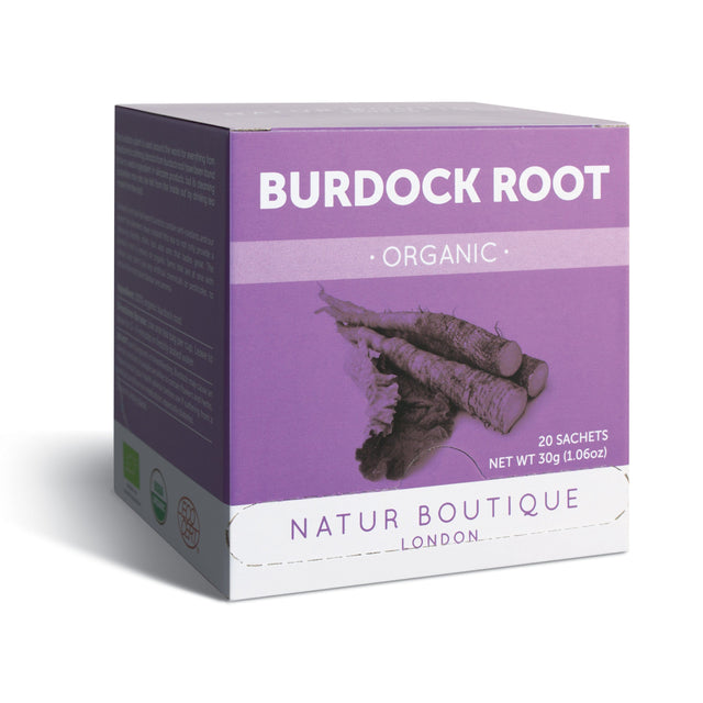 A close-up of a tea infuser with brewed organic burdock root tea in a clear glass cup, highlighting the rich amber colour, surrounded by fresh burdock roots and green tea leaves on a rustic wooden surface, illustrating the natural and organic qualities of Natur Boutique Organic Burdock Root Tea, 20 Sachets